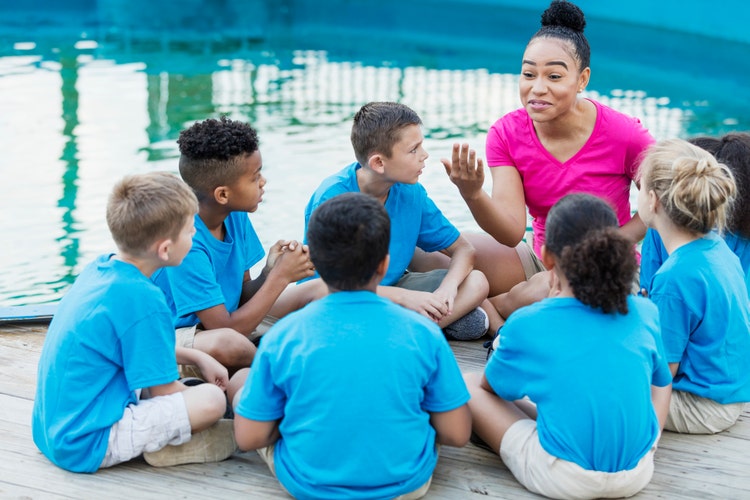 A group of multi-ethnic children on a field trip to a marine park. They are sitting in a circle by a pool, listening intently to a young African-American woman telling a story.