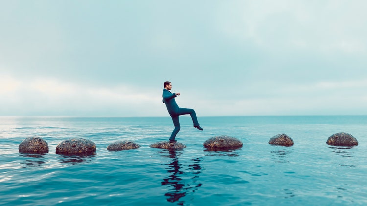 Man walks carefully on a path of small rocks in the middle of the sea. He looks focused and moves in the right direction. Concept of success and avoiding problems on the way of a journey or career path.