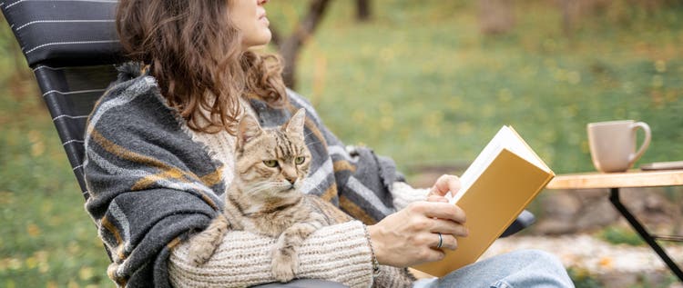 Young woman sitting in the autumn garden with a pet gray cat in her hands reading literature