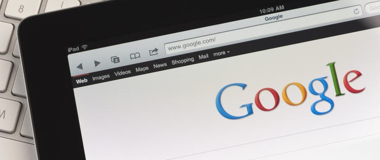 Close up of an Apple Ipad and on a desk showing the Google search engine home screen. There is also a keyboard and some documents on the desk, the documents show charts and graphs. This scene depicts ipad in a working environment.