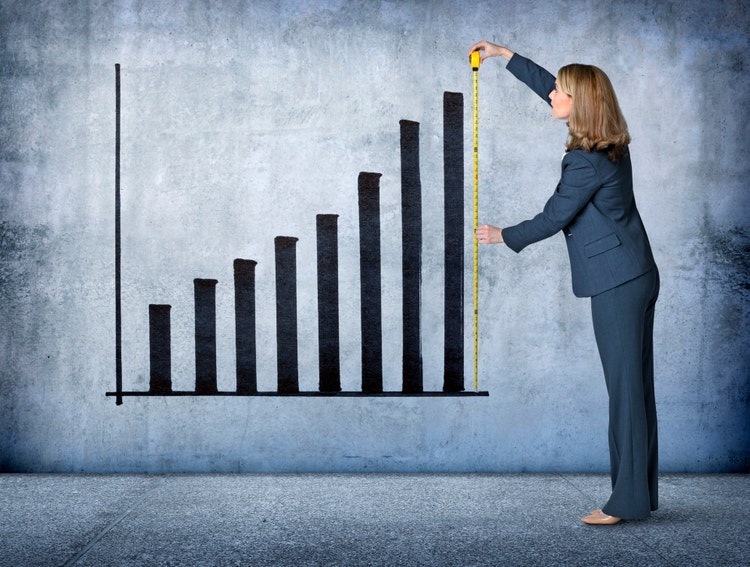 A businesswoman uses a tape measure to measure the growth of an upward pointing graph projected on a concrete wall.