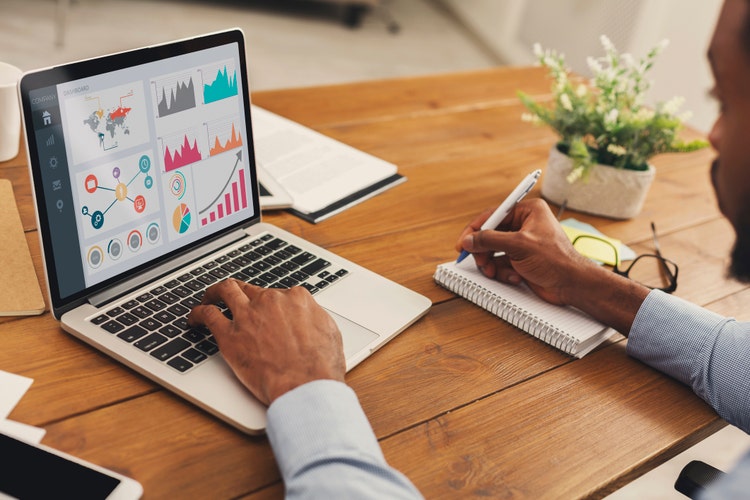 Close up back view of black business person analyzing graph on laptop and writing in notepad