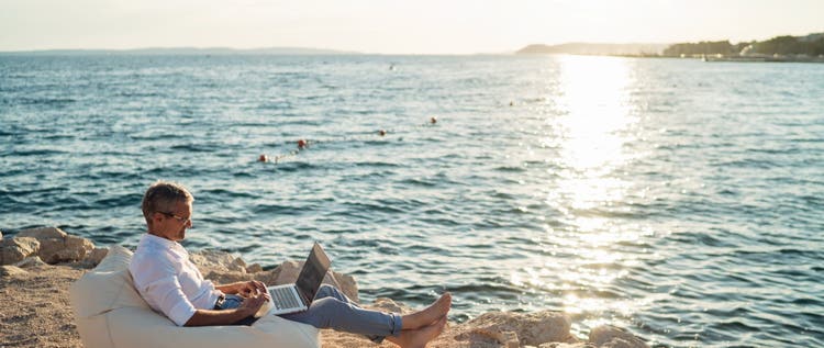 A man working on a laptop on a beach while sitting in a beanbag chair.