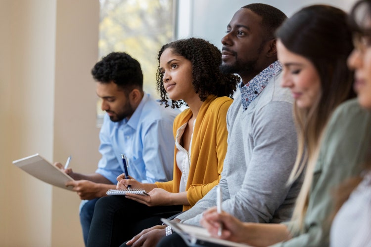A group of business people sit in a row in a training class. They look at an unseen speaker as they concentrate on his lecture.