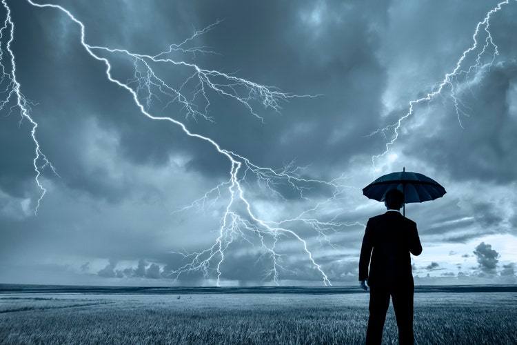 A rear view of a businessman in a suit, with an upheld umbrella, standing in a large field during a thunderstorm. Lightning is seen descending from a gray and cloudy sky.