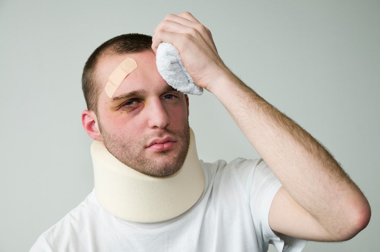 A beat-up young man in white wearing a neck brace and holding an icepack to his head