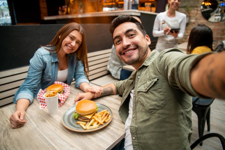 Happy Latin American couple taking a selfie while eating burgers at a restaurant to post on their social media - lifestyle concepts