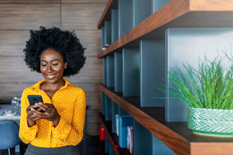 Quick message to friend. Attractive young African woman holding smart phone and looking at it while standing indoors.