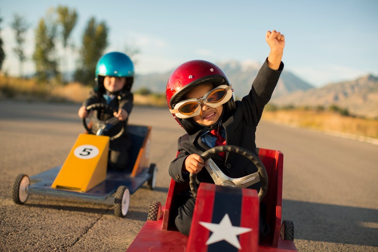 two young children racing in pretend racecars. The winner is raising a fist in the air.