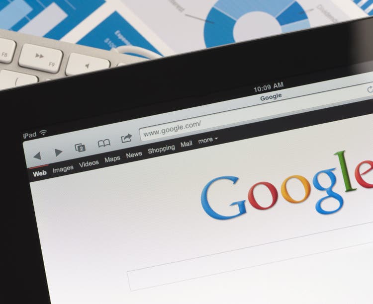 Close up of an Apple Ipad and on a desk showing the Google search engine home screen. There is also a keyboard and some documents on the desk, the documents show charts and graphs. This scene depicts ipad in a working environment.