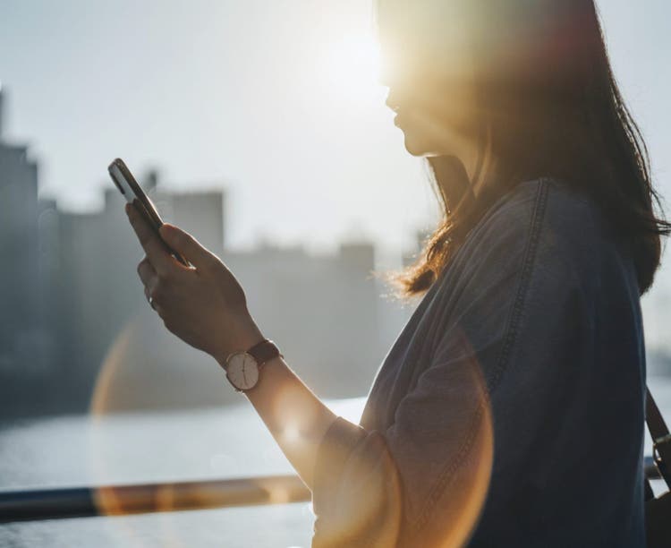 Person in silhouette looking at a smartphone outdoors, backlit by the sun with golden lens flare.