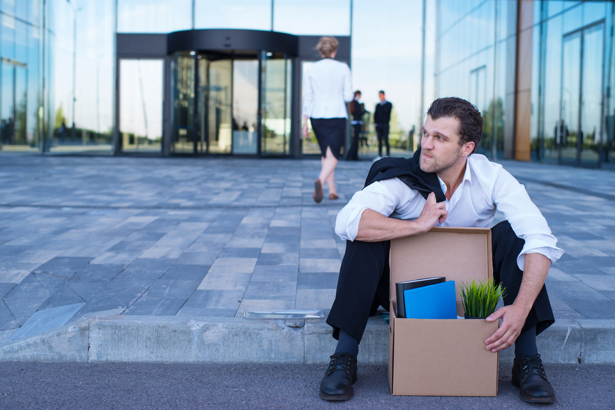 Fired business man sitting frustrated and upset on the street near office building with box of his belongings. 