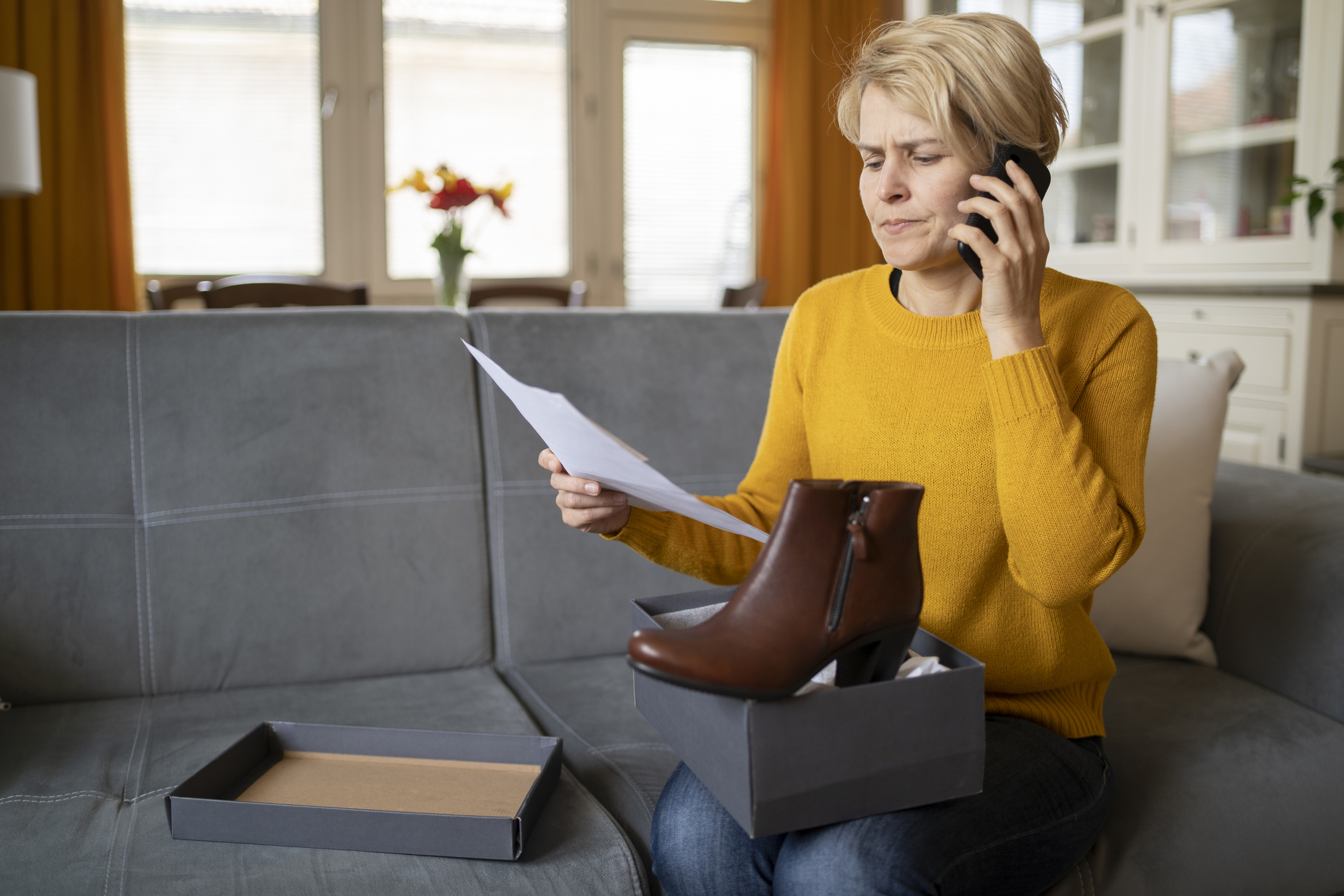 An dissatisfied customer is looking at the delivery note from her online shopping order, using her mobile phone, the new purchase of boots on in the box on her knee while she sits in the living room on her sofa