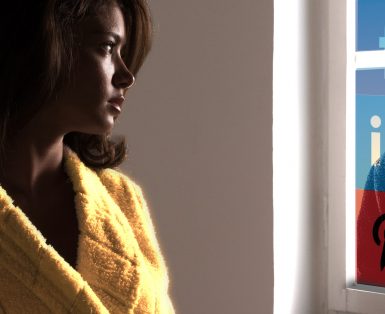 A woman in a yellow bathrobe looking through a window made up of social media icons to the shadow of a man looking in.