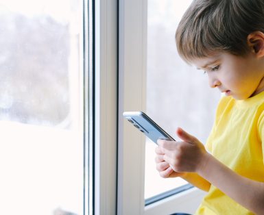 Little toddler boy is holding the smartphone at home. He is wearing bright yellow t-shirt. Playing video games like a social problem