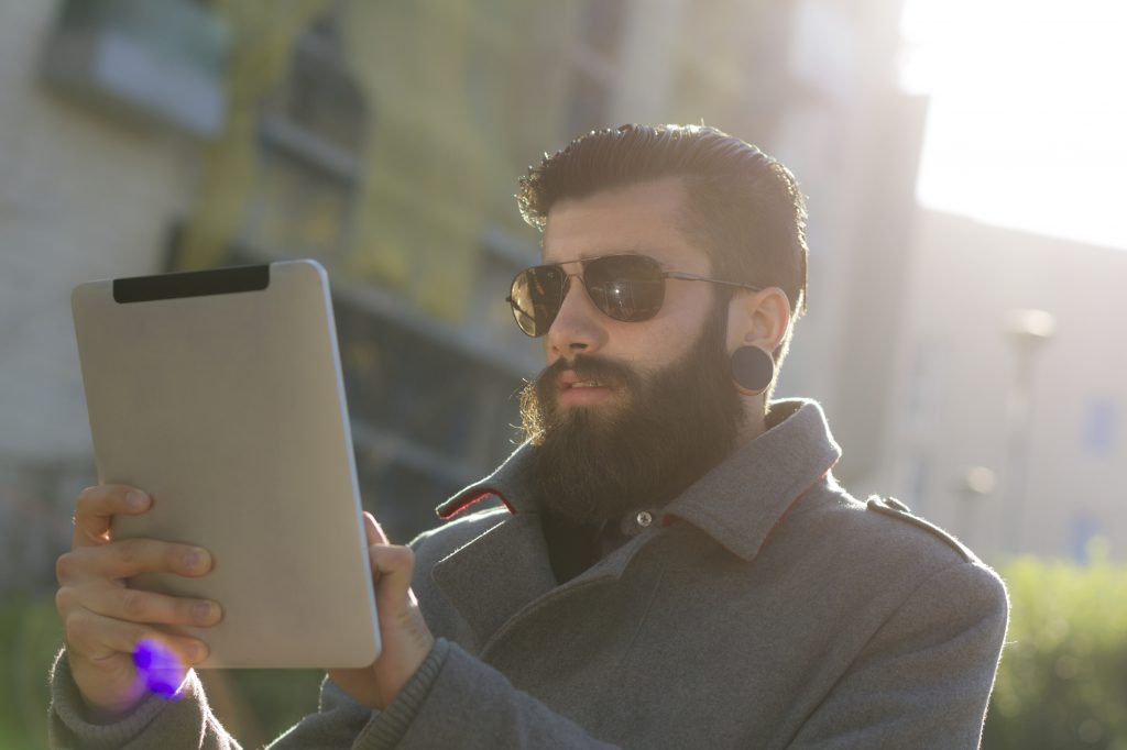 Young man (hispter style) with big earring and full beard, wearing sunglasses using a digital tablet outdoor. Sunlight and flares on picture.