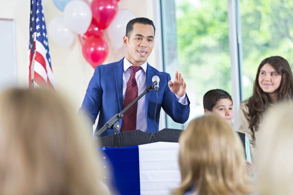 Mid adult Hispanic man is giving speech and answering questions while running for local city government political office. His children are standing on stage next to him in support.