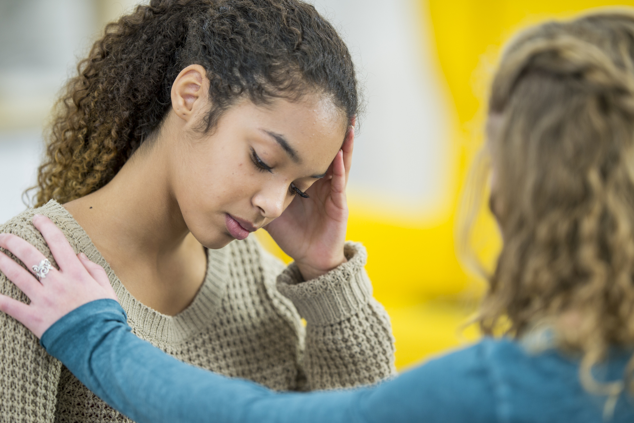 Two young women are indoors in a bedroom. They are wearing casual clothing. One woman looks sad, and she is being comforted by her friend.