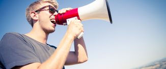 Young caucasian man screaming with a megaphone on the top of a hill during a sunny day.