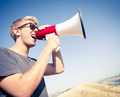 Young caucasian man screaming with a megaphone on the top of a hill during a sunny day.