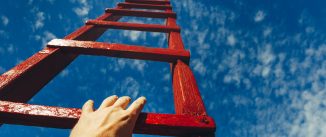 A male hand holds onto the crossbar of a red wooden staircase leading to the blue sky