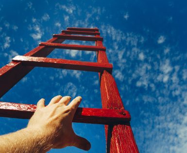 A male hand holds onto the crossbar of a red wooden staircase leading to the blue sky