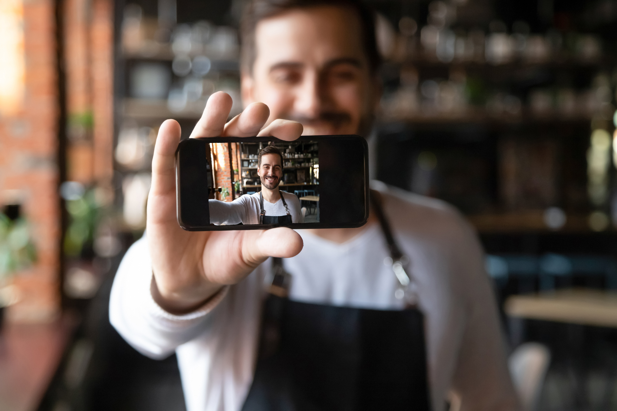 Happy man wearing apron holds smartphone with camera on, waiter or cafe owner make selfie photo focus on device with self portrait image on mobile screen, share life in social media having fun concept