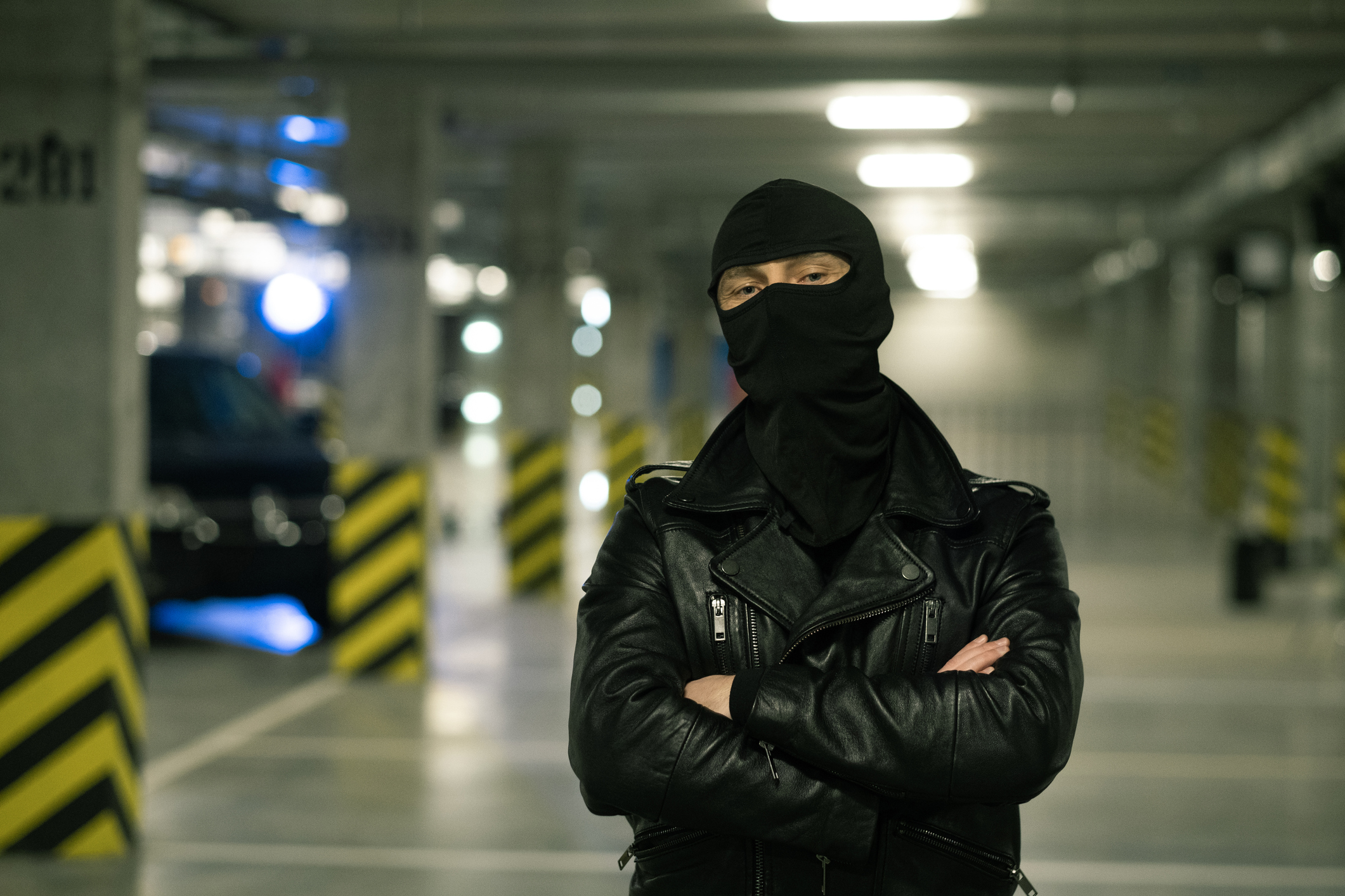 Contemporary criminal in black leather jacket and balaclava on head crossing arms by chest with parking area on background