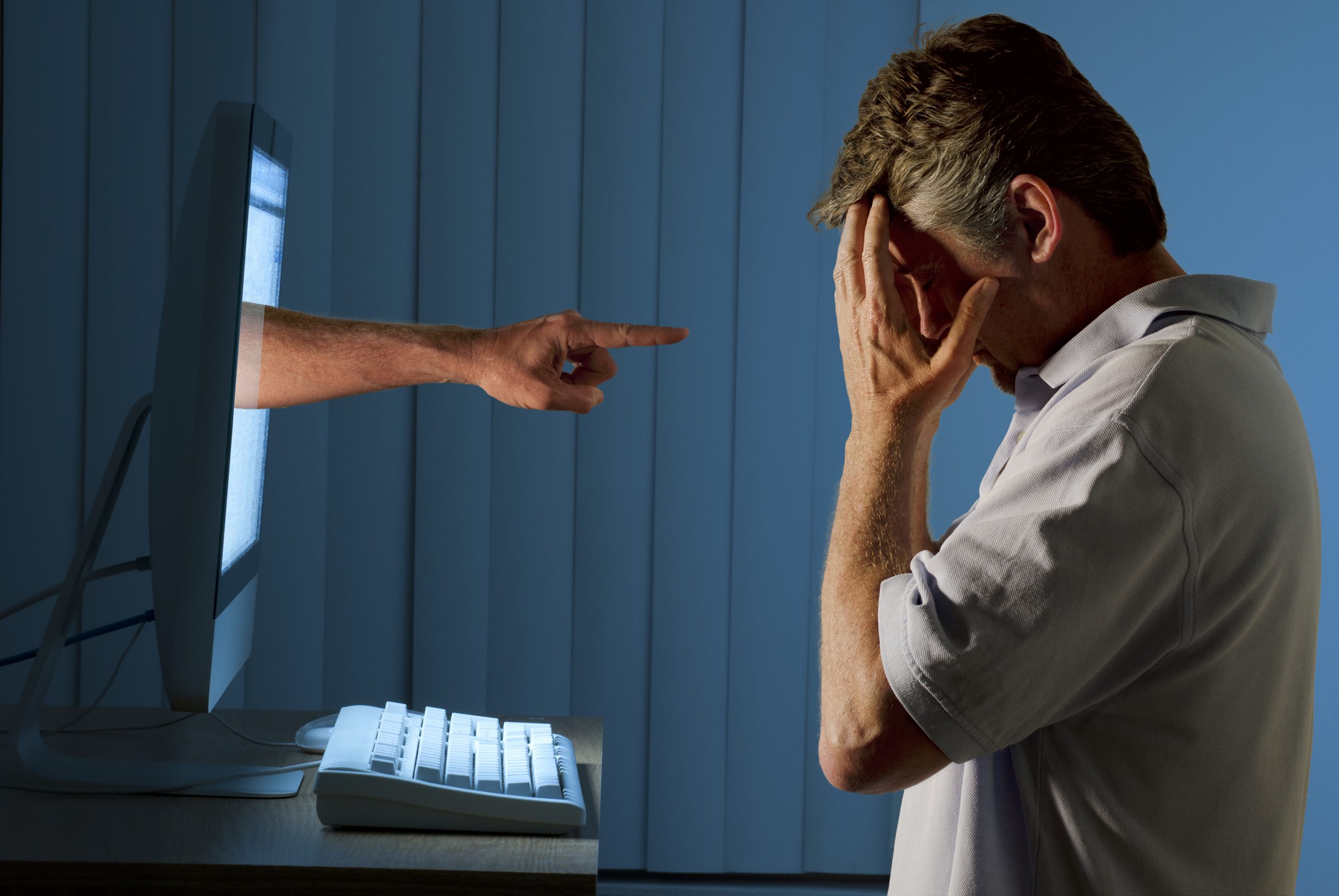 Severely distraught young man sitting in front of a computer with a judgmental hand pointing at him from within the computer monitor which shows the man being either computer bullying bullied or Facebook social media stalking stalked.