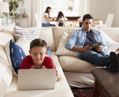 Pre-teen boy lying on sofa using laptop, dad sitting with tablet, mum and sister in the background