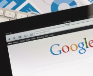 Close up of an Apple Ipad and on a desk showing the Google search engine home screen. There is also a keyboard and some documents on the desk, the documents show charts and graphs. This scene depicts ipad in a working environment.