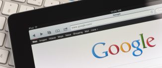 Close up of an Apple Ipad and on a desk showing the Google search engine home screen. There is also a keyboard and some documents on the desk, the documents show charts and graphs. This scene depicts ipad in a working environment.