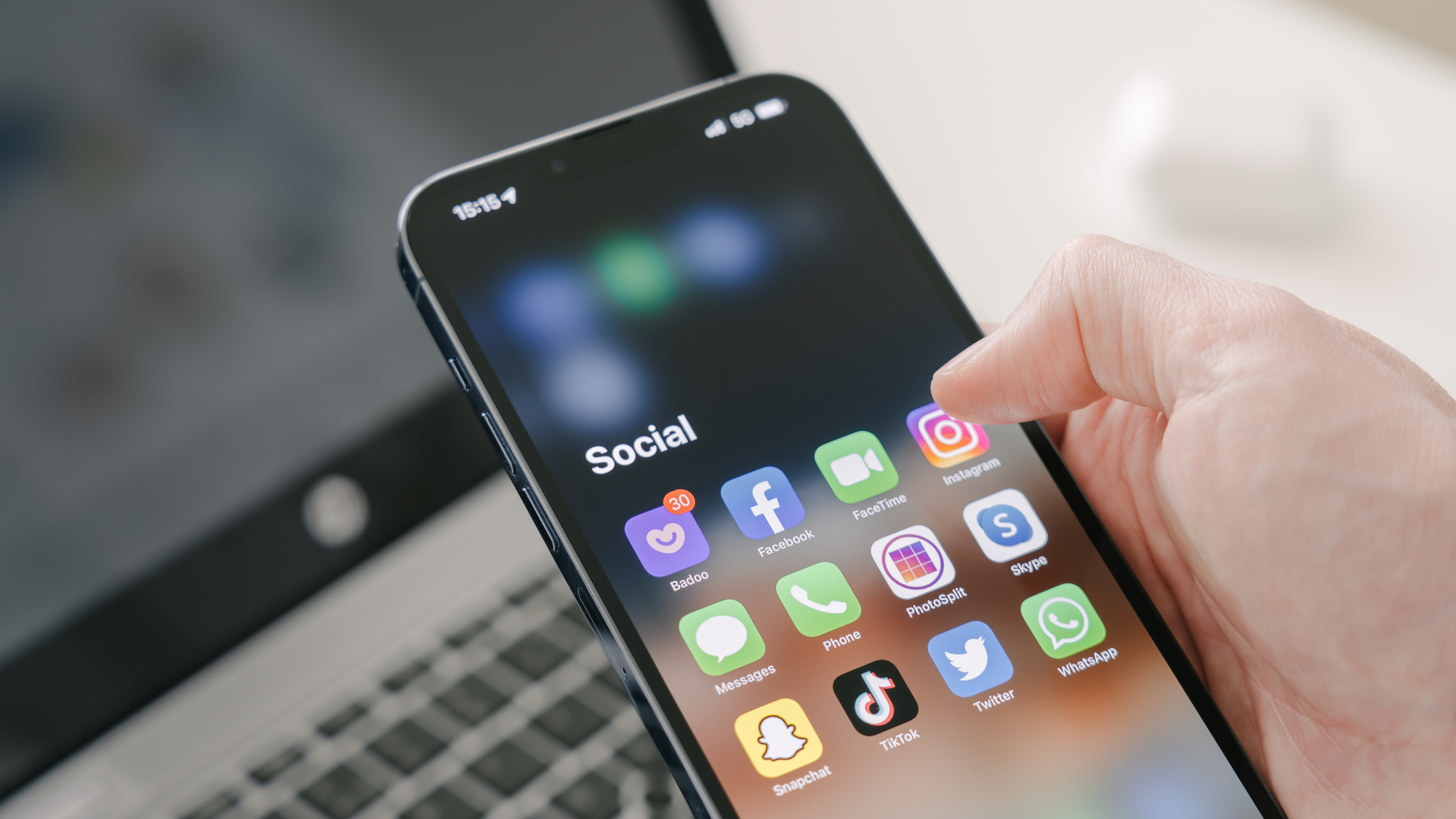 A man's hand holds a Iphone 13 Pro max with social media apps, against the background of a laptop. Blurred background. Selective focus.