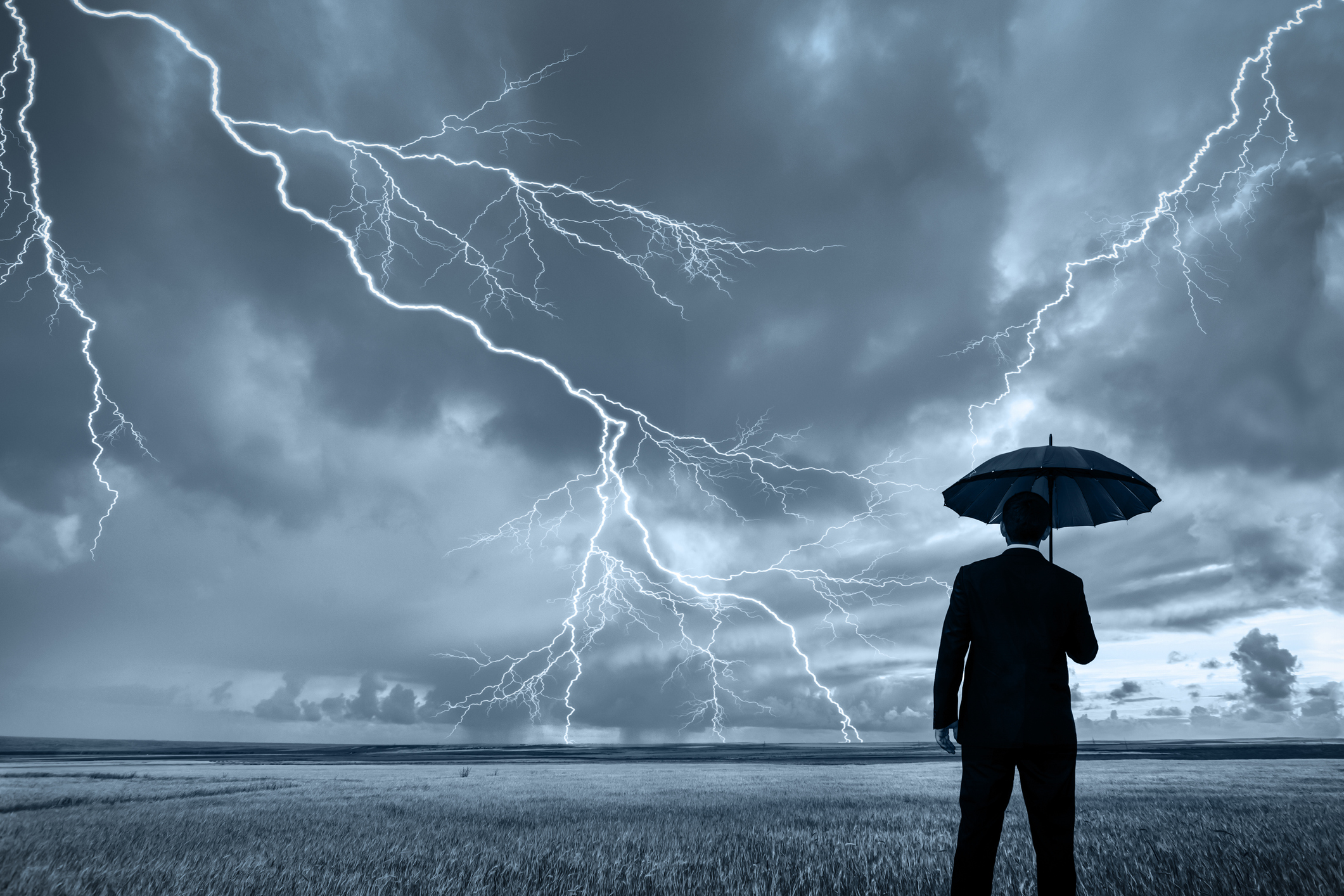 A rear view of a businessman in a suit, with an upheld umbrella, standing in a large field during a thunderstorm. Lightning is seen descending from a gray and cloudy sky.
