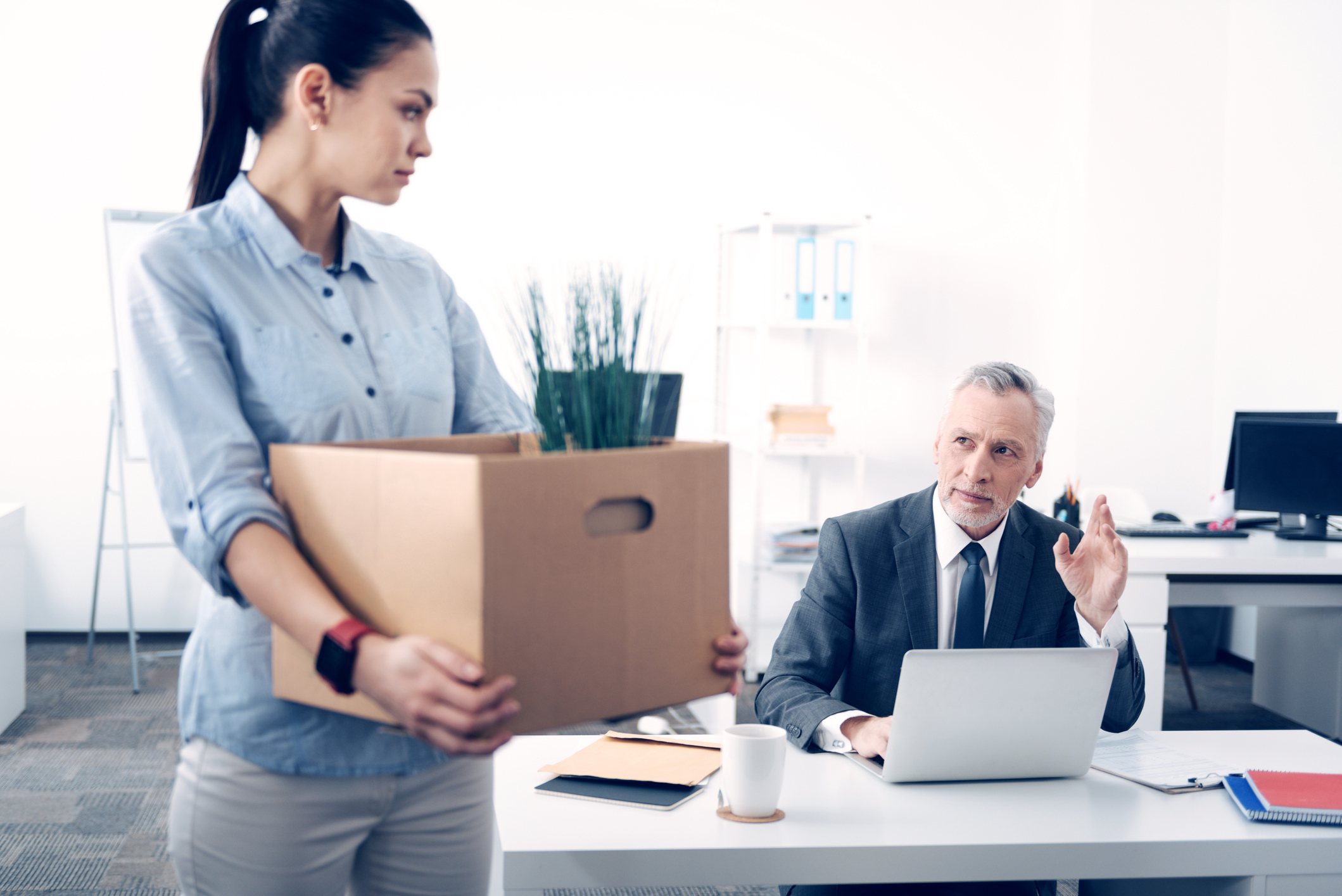 Saying goodbye. Selective focus on a mature boss looking at his serious female employee with a slight on his face and waving a farewell while a young lady leaving her work with a paper box in