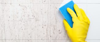 Closeup of a hand in a yellow rubber glove cleaning a dirty bathroom surface with a blue sponge.