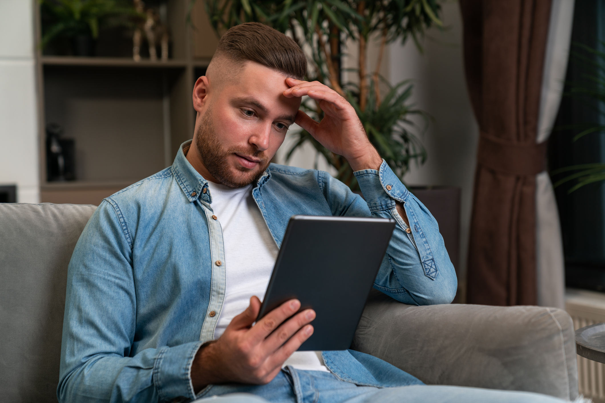 Pensive man sitting on sofa with tablet and hand on head, virtual meeting and video conference. Concept of distance work and education