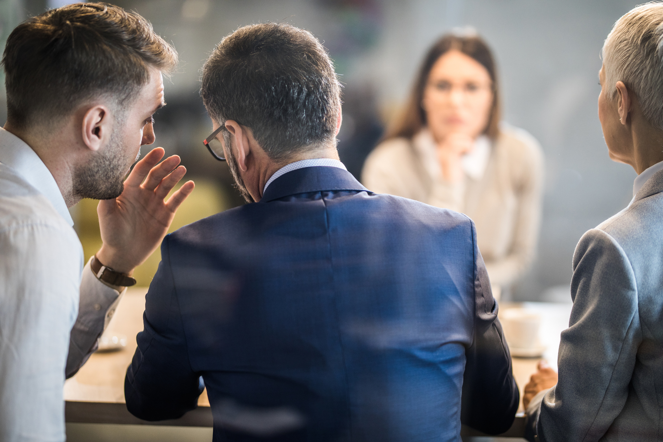 Male member of human resource team whispering to his colleague during a job interview in the office. The view is through glass.