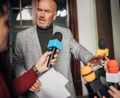 Group of people, male politician confronted by journalists with microphones.