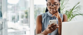 Young black businesswoman using a cellphone while working on a laptop in an office alone