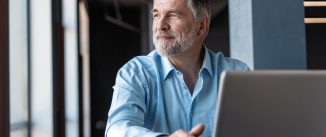 A middle-aged man with a beard is seated at a table in front of an open laptop, looking out contentedly of a window.