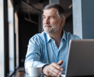 A middle-aged man with a beard is seated at a table in front of an open laptop, looking out contentedly of a window.